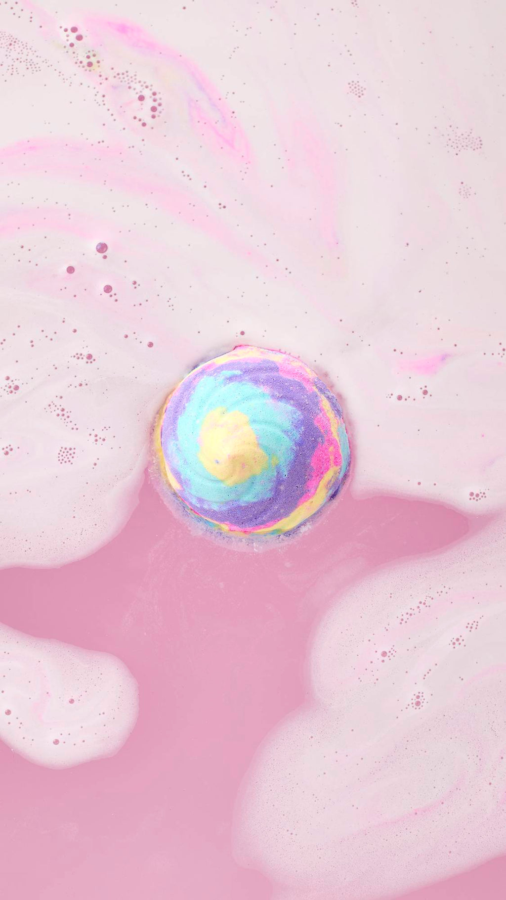 Top-down view of the Unicorn Poop Bath Bomb floating in a pool of milky pink bathwater, releasing soft swirls of pastel yellow, blue, and purple foam.