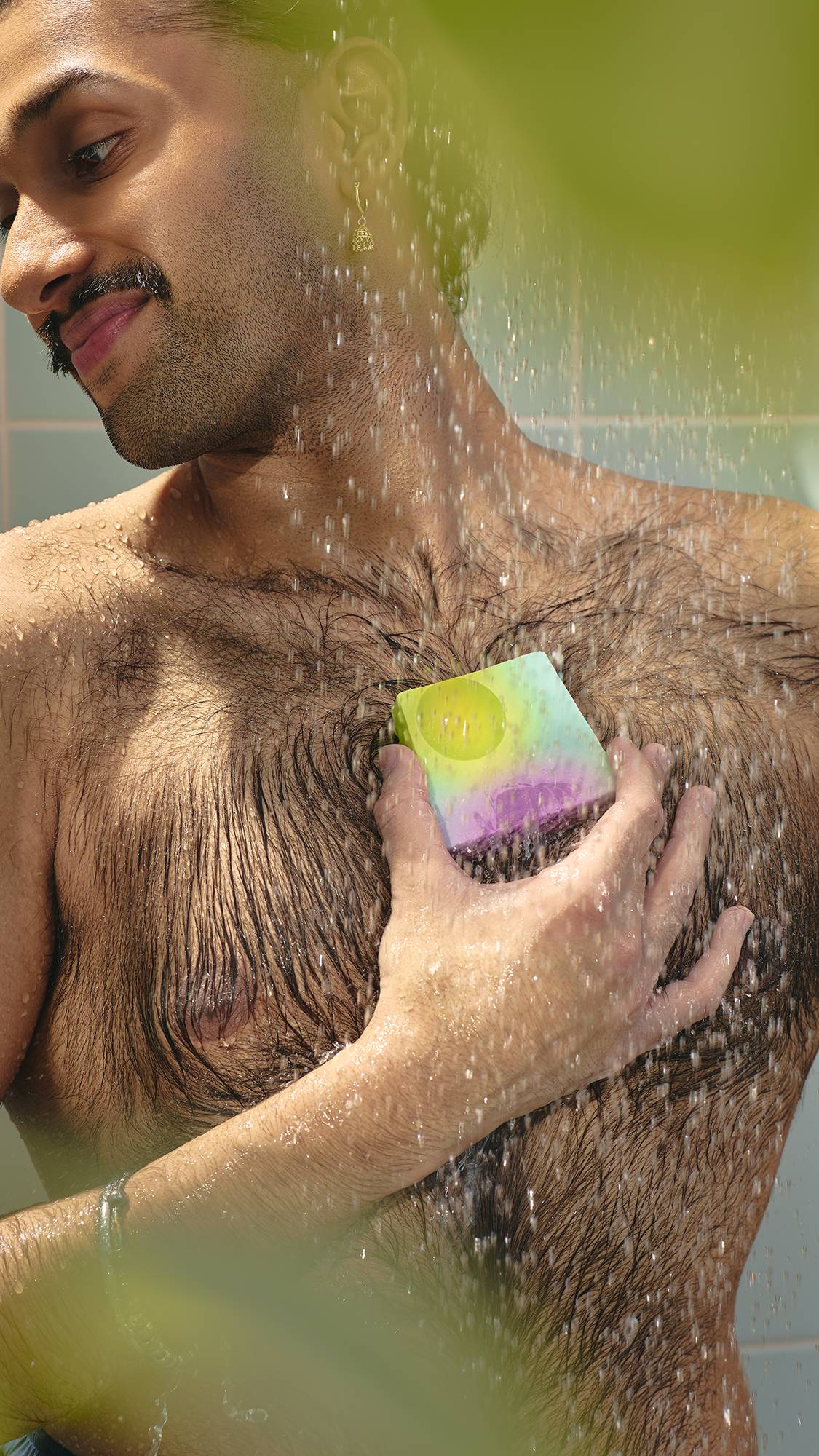 The image shows a model with dark hair in a bun looking down as they lather their chest with a slice of marbled purple and green "To The Moon and Back" soap in the shower. The water creates a fine mist around
