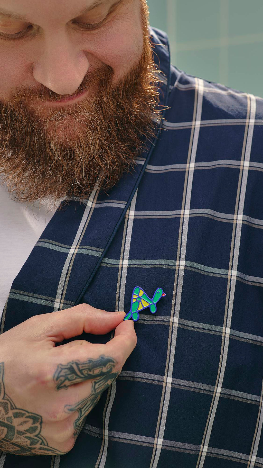 A model with a thick beard and hand tattoos smiles as they pin a small, colourful turtle enamel badge, from the "Turtley Awesome" greeting card, onto the lapel of his navy blue and white checked dressing gown.