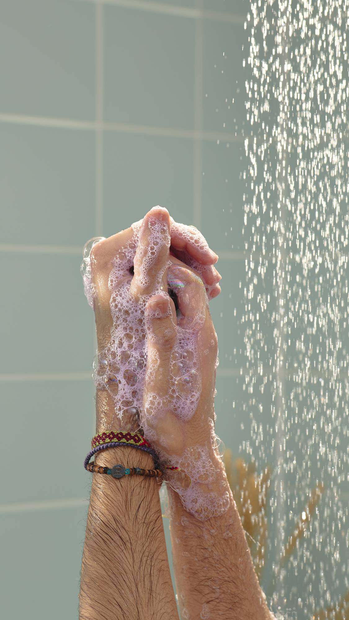 A close-up image of a model's hands covered in thick, pale purple "Stormy Weather" shower gel as they lather in the shower. A stream of falling water is visible on the right against light green tiles.