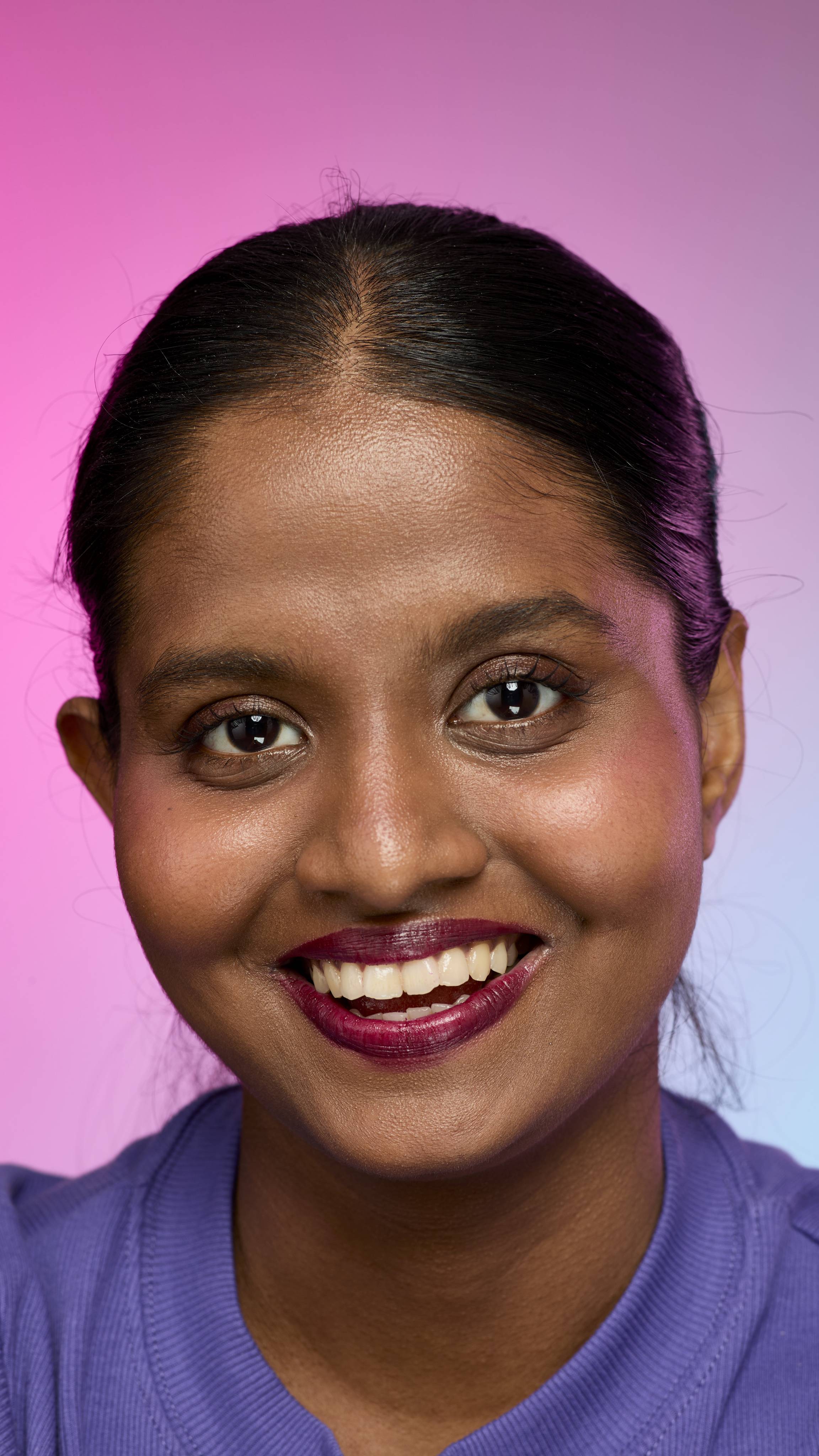 A smiling person with dark hair and skin, wearing a purple top, is shown from the chest up. They are wearing the Lush, deep plum Tartlet lip cream on a pink and purple gradient background.