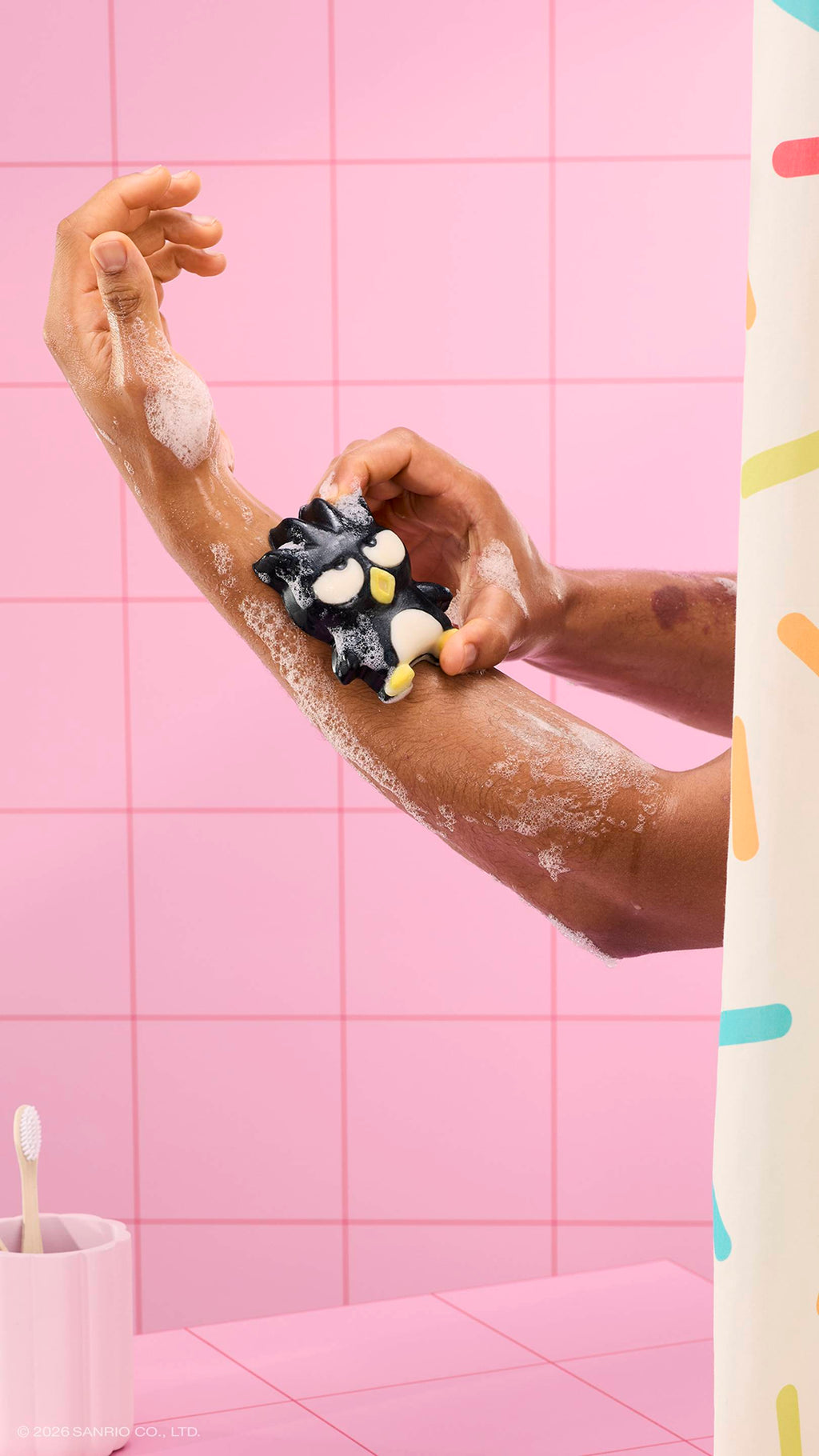 A person uses a black penguin-shaped "Bad Badtz-Maru" soap character to lather their arm in a pink tiled bathroom. The soap is covered in white bubbles, and a sprinkle-patterned shower curtain is visible on the right.