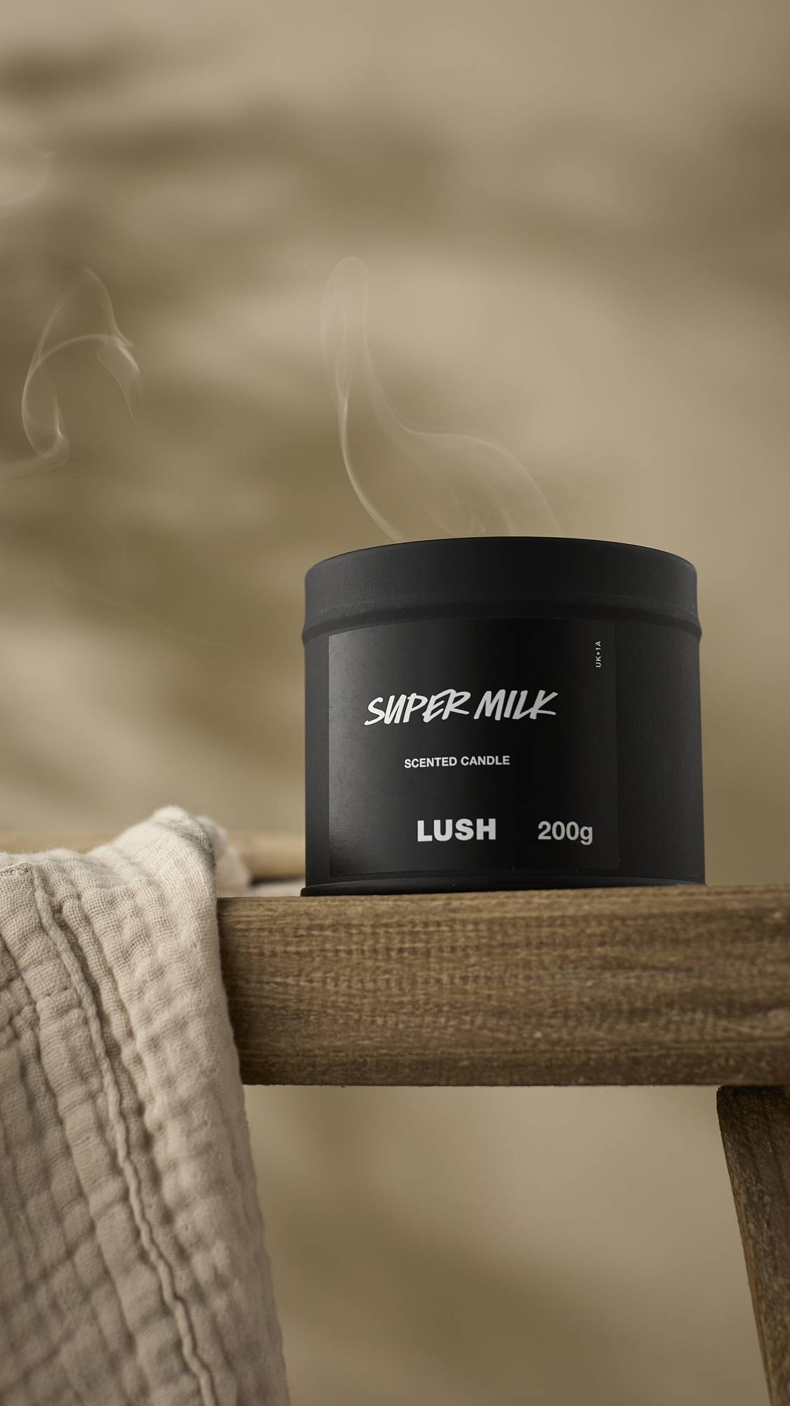 The matte black Super Milk Scented Candle is shown with its lid on, sitting on the edge of a rustic wooden frame or stool. A wisp of white smoke rises from the tin, and a light-coloured, textured cloth hangs in the foreground.