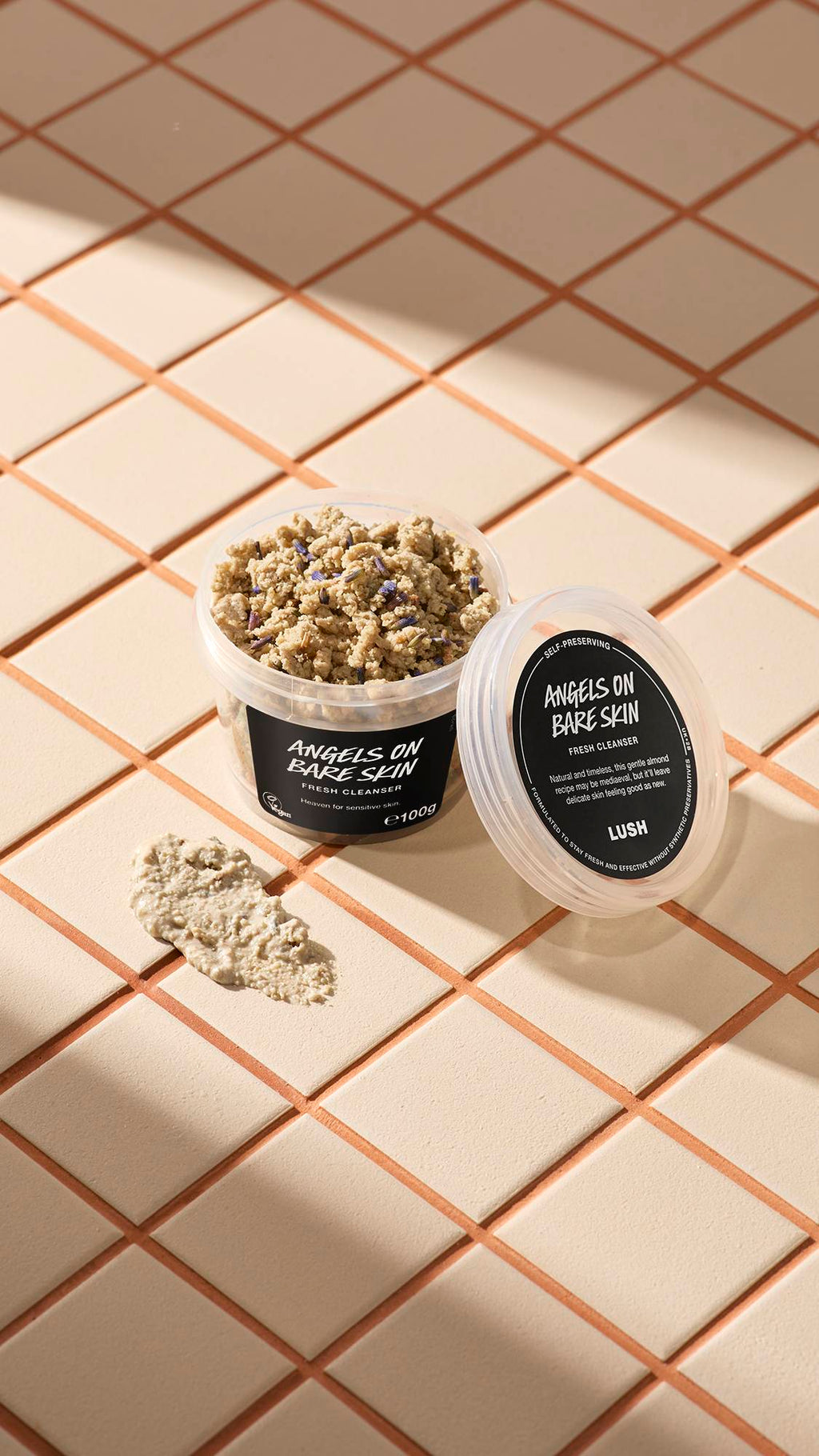 An open clear pot of Lush Angels on Bare Skin Fresh Cleanser, showing a crumbly lavender-flecked paste, sits on beige tiles. Its lid rests alongside it next to a smear of the creamy mixture, illuminated by soft diagonal sunlight.