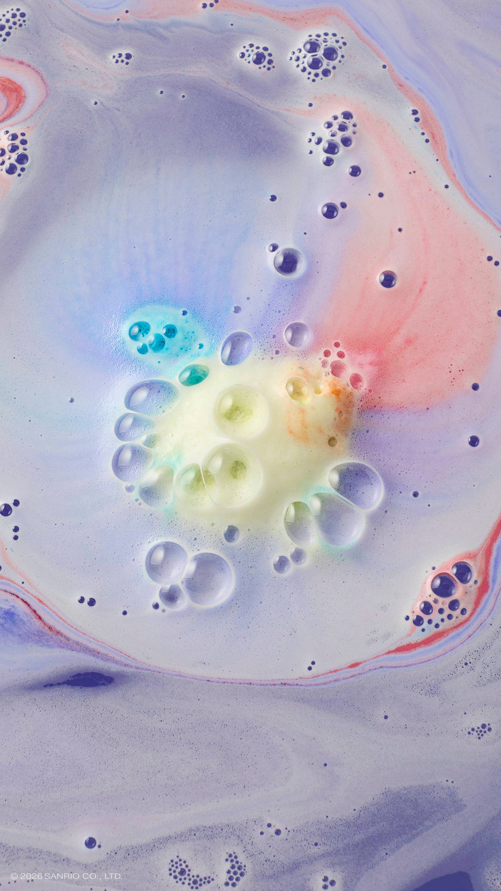 An aerial shot of lavender bath water swirling with multi-coloured foam. In the centre, a cluster of yellow and blue bubbles marks where the "U.SA.HA.NA" bath bomb has dissolved, with soft streaks of pink and purple radiating across the surface.