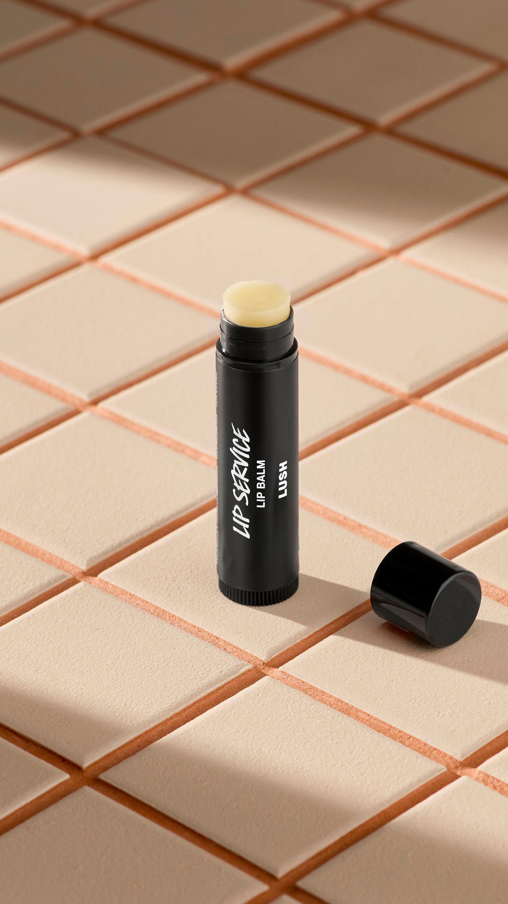 A black tube of Lush 'Lip Service' lip balm stands upright on light-coloured square tiles. The black cap is off, lying next to the base, revealing the pale, creamy balm inside. Soft shadows fall across the tiled surface.