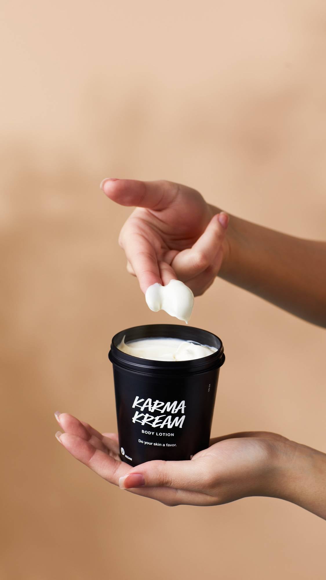 Hands hold a black pot of Lush Karma Kream body lotion against a warm beige background. One hand holds the open tub, while fingers scoop out a dollop of the rich, white cream.