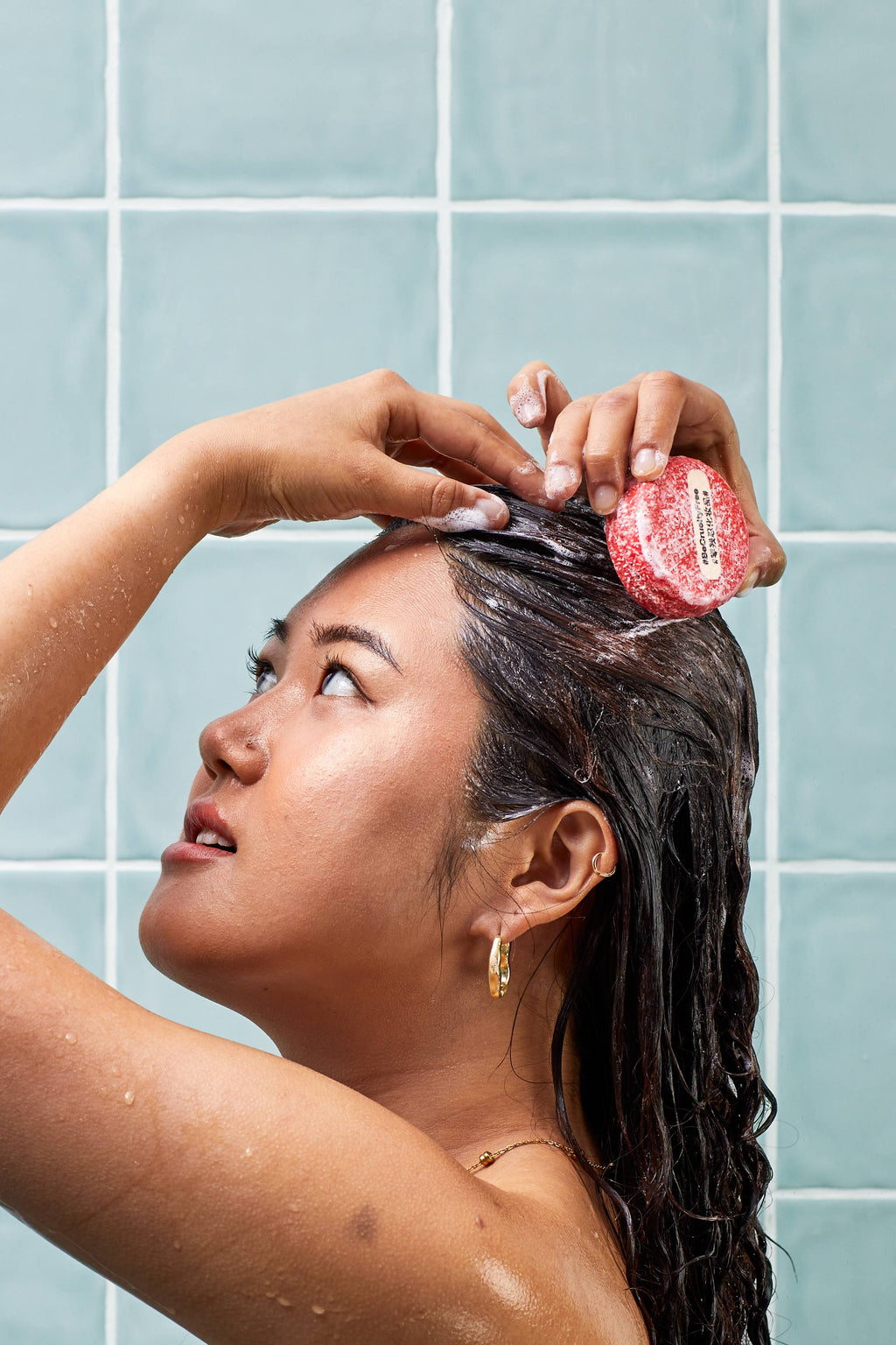A side profile of a person lathering their wet, dark hair with a textured red circular "NEW" shampoo bar. Their hands are raised to their head, working up a light foam against a background of pale blue-green wall tiles.