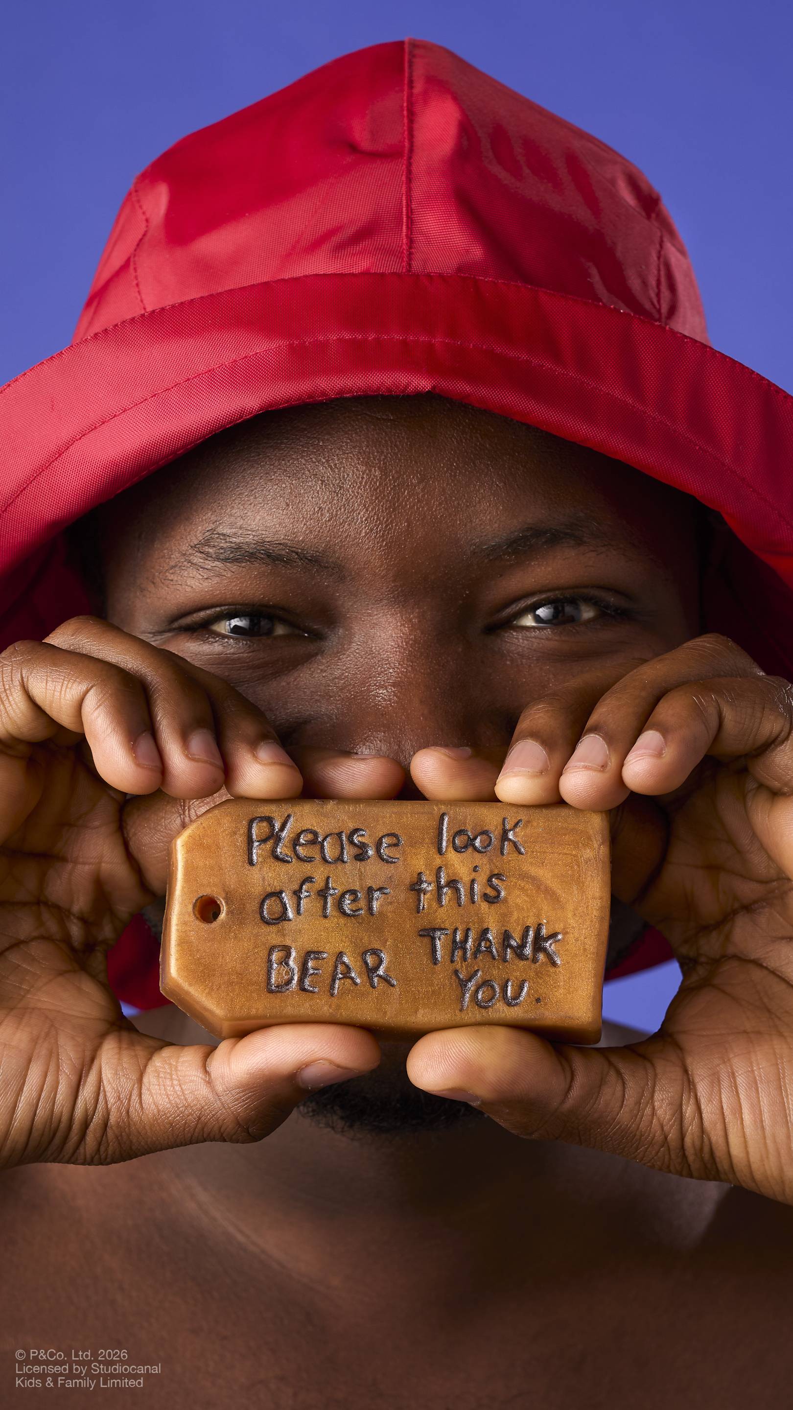 A model wearing a red rain hat holds a brown, luggage-tag-shaped soap in front of their face. The soap features the embossed, handwritten-style message 'Please look after this bear, thank you'. On a solid blue background.