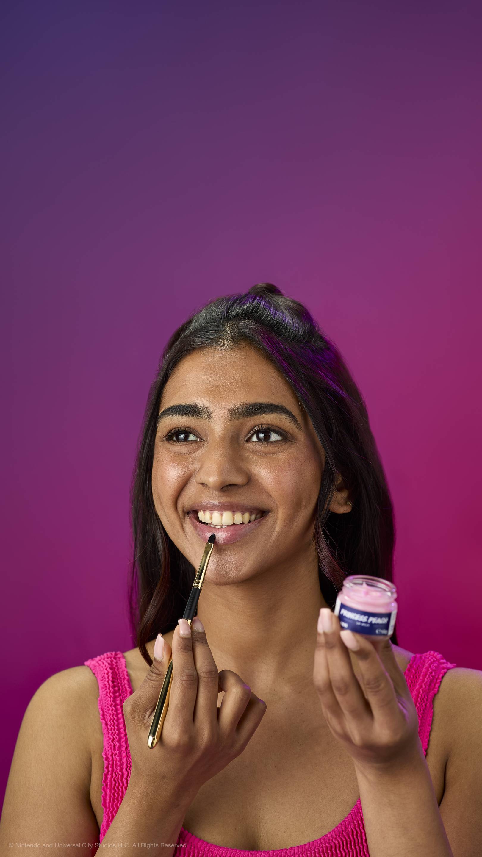 A woman in a pink top smiles while applying pink Princess Peach lip jelly with a gold brush. She holds the open glass pot against a purple-magenta gradient background.