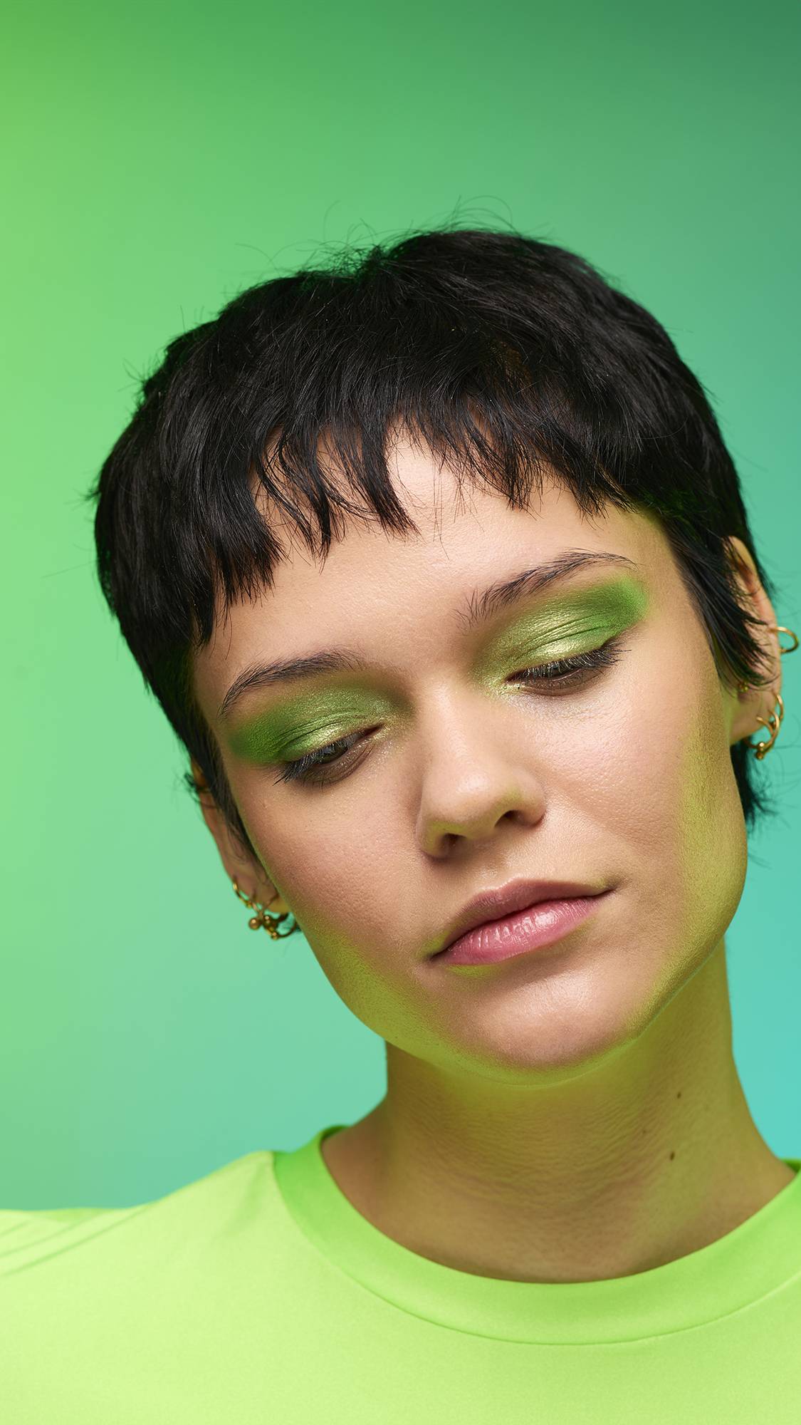 The image shows a close-up of the model's face on a pale blue-to-green gradient background. The model's make-up is done with the focal point being a subtle, jade-green, shimmering eyeshadow made by the Aurora Glitter Crumble.