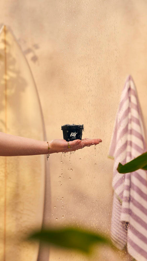 An outstretched hand holds a small black Lush pot labelled 'Big shampoo' under a spray of falling water. In the warm, neutral background, a surfboard rests on the left and a striped towel hangs on the right.