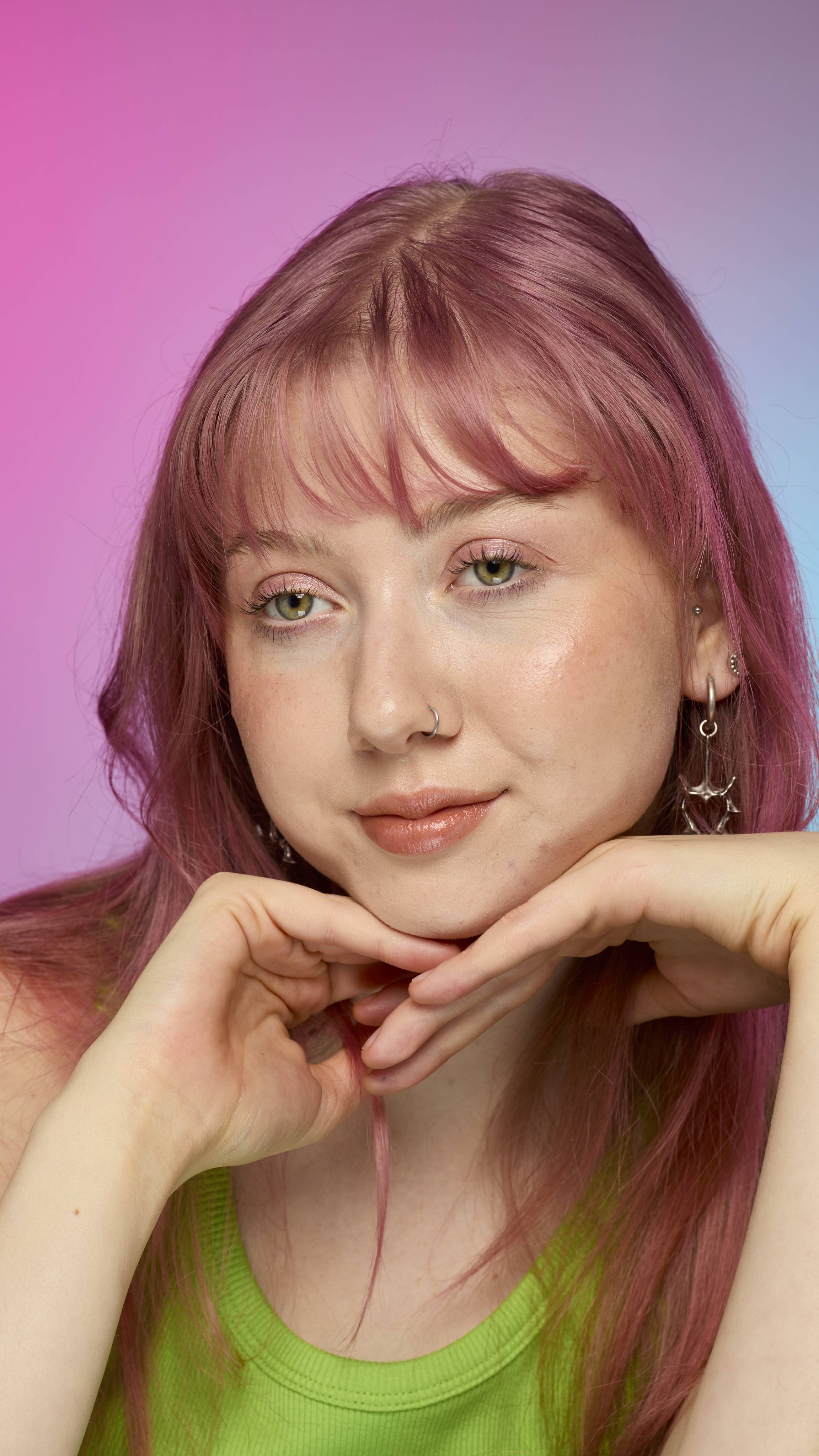 The image shows a fair-skinned model on a pink and blue gradient background as they rest their chin on their hands. They have pink hair and subtle shimmering lips from using the Crackled Copper lip cream.