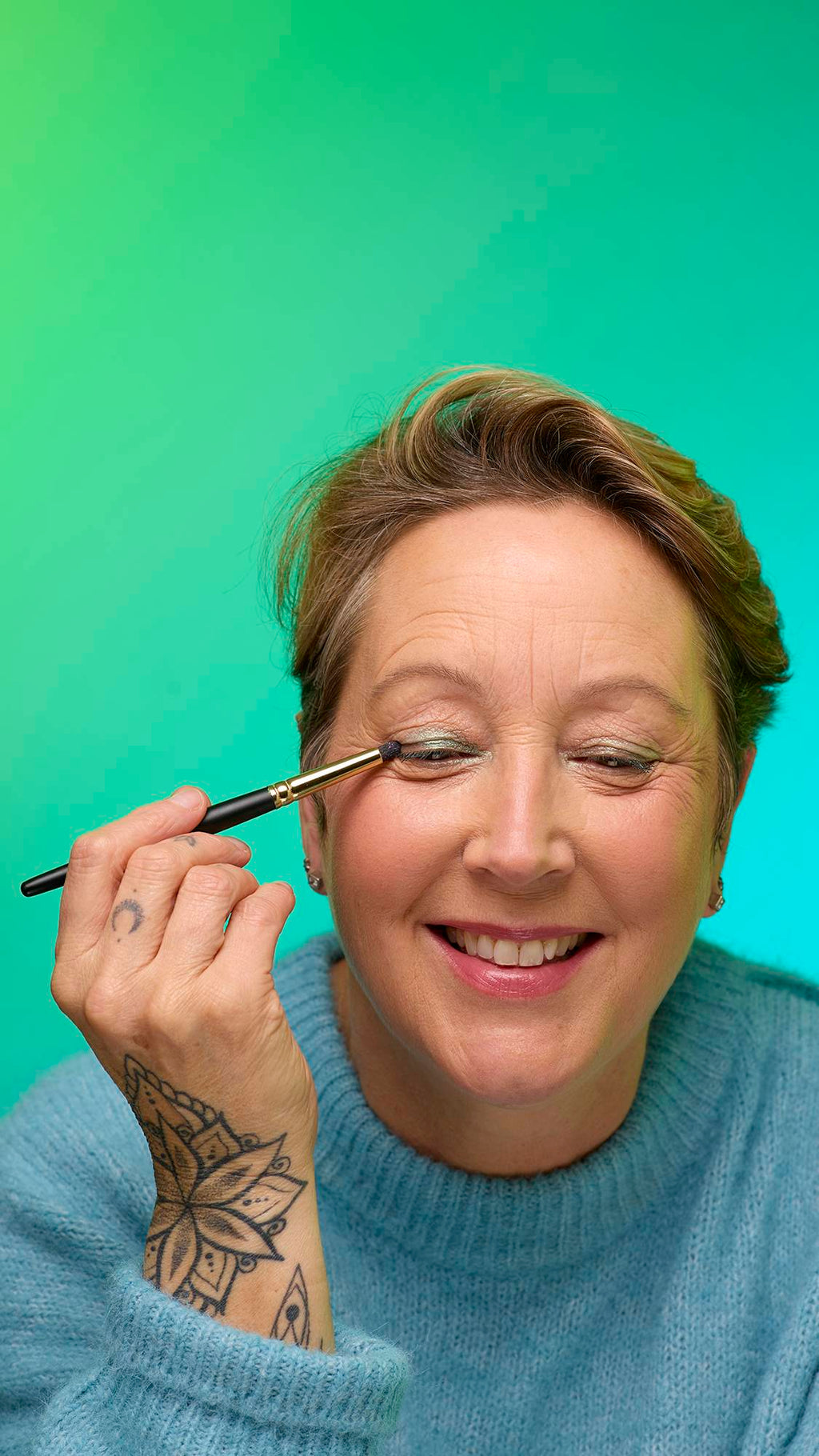 The image shows a close-up of the model's face on a pale blue-to-green gradient background. They are holding a thin make-up brush to apply a subtle, deep-sage-green, shimmering eyeshadow of the Meteor Glitter Crumble.