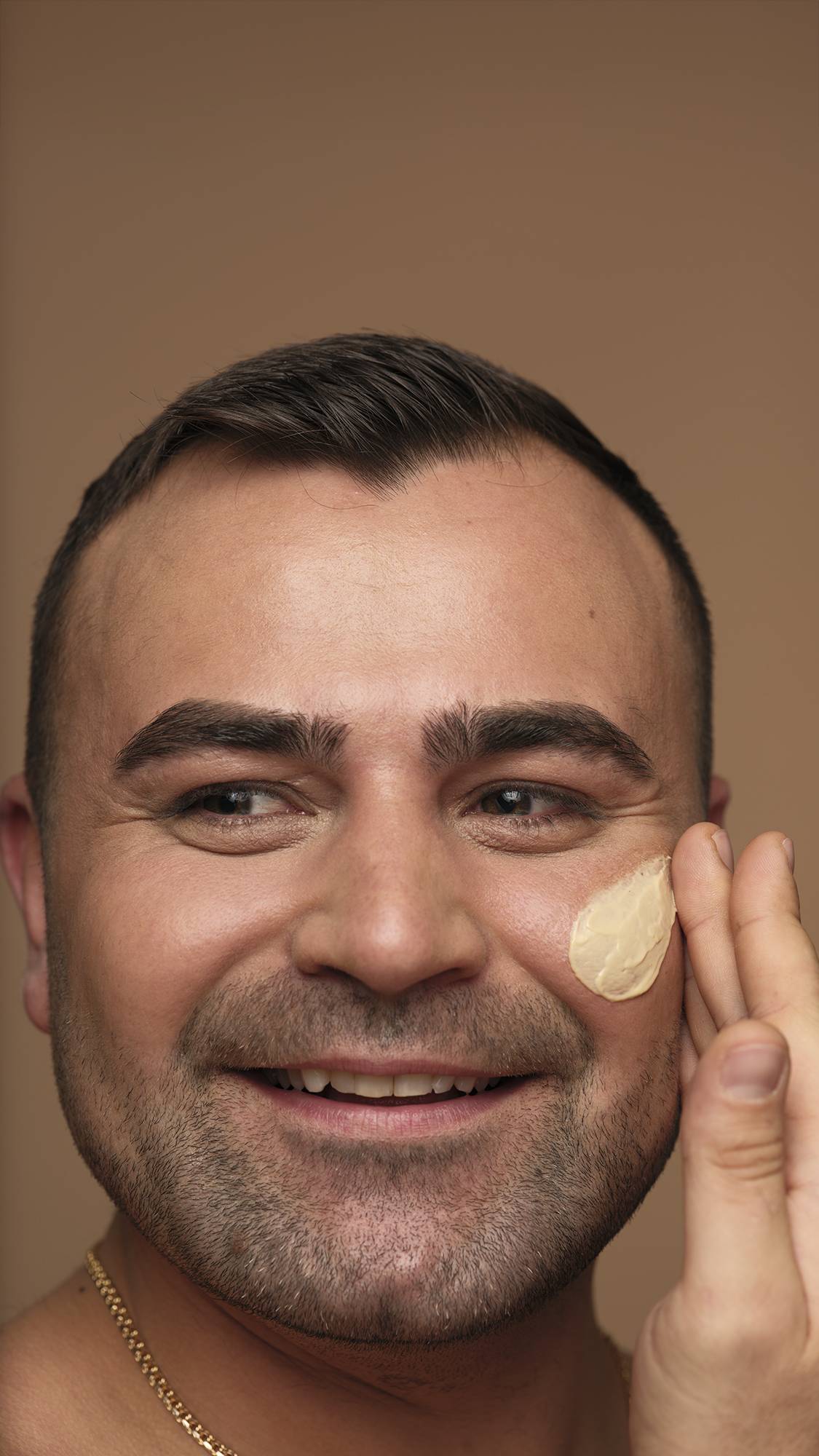 A close-up photograph of a smiling person with short dark hair and facial hair applying Skin Drink moisturiser to their cheek with their fingers against a neutral background.