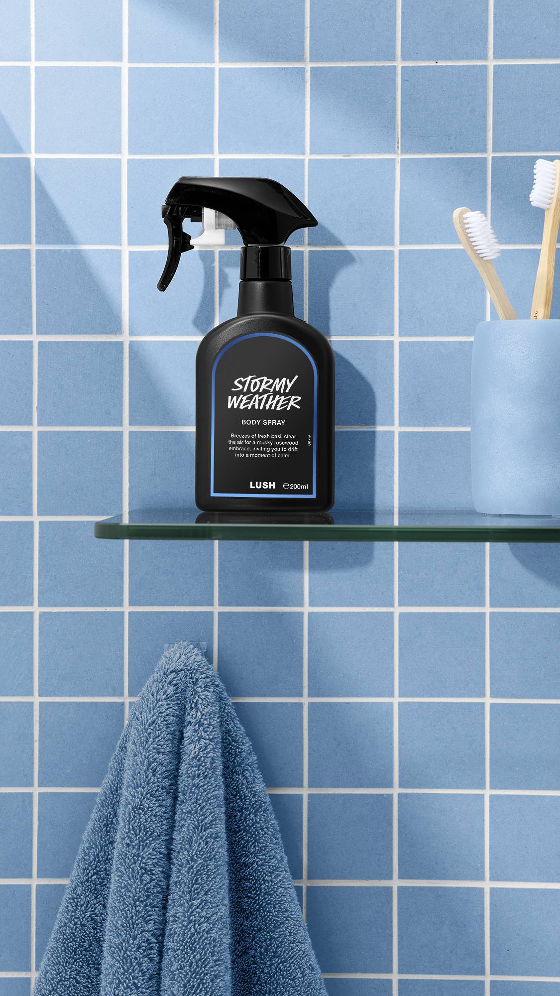 The image shows a bottle of LUSH 'Stormy Weather' body spray on a glass shelf. A blue towel hangs below, with a blue cup and toothbrushes nearby, against a bright, tiled, sky-blue bathroom wall.
