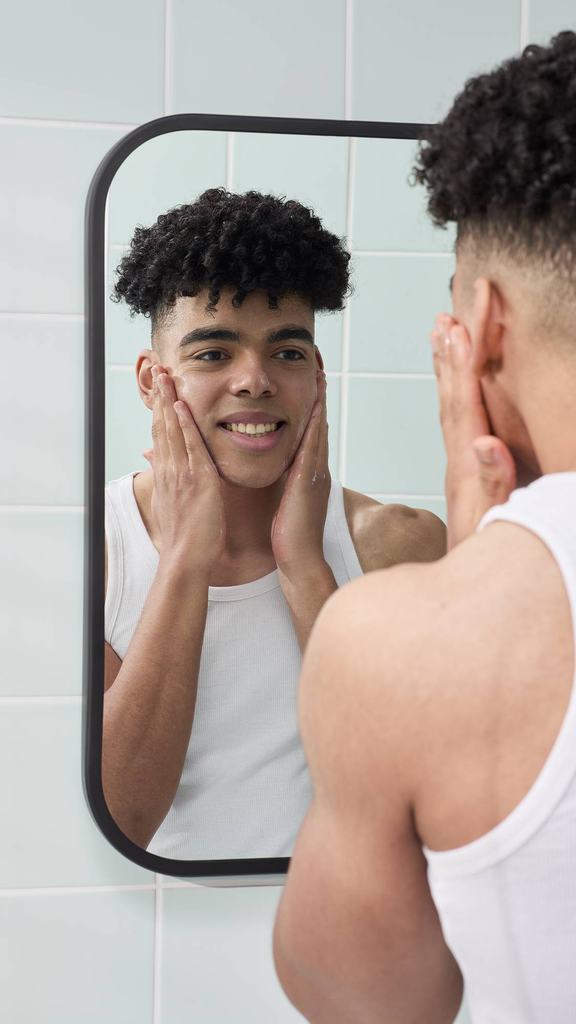 A reflection in a bathroom mirror shows a smiling model with short, curly dark hair applying a lather to their face, created by the Strawberry Picking facial gummy. They are wearing a white vest against a background of pale green wall tiles.