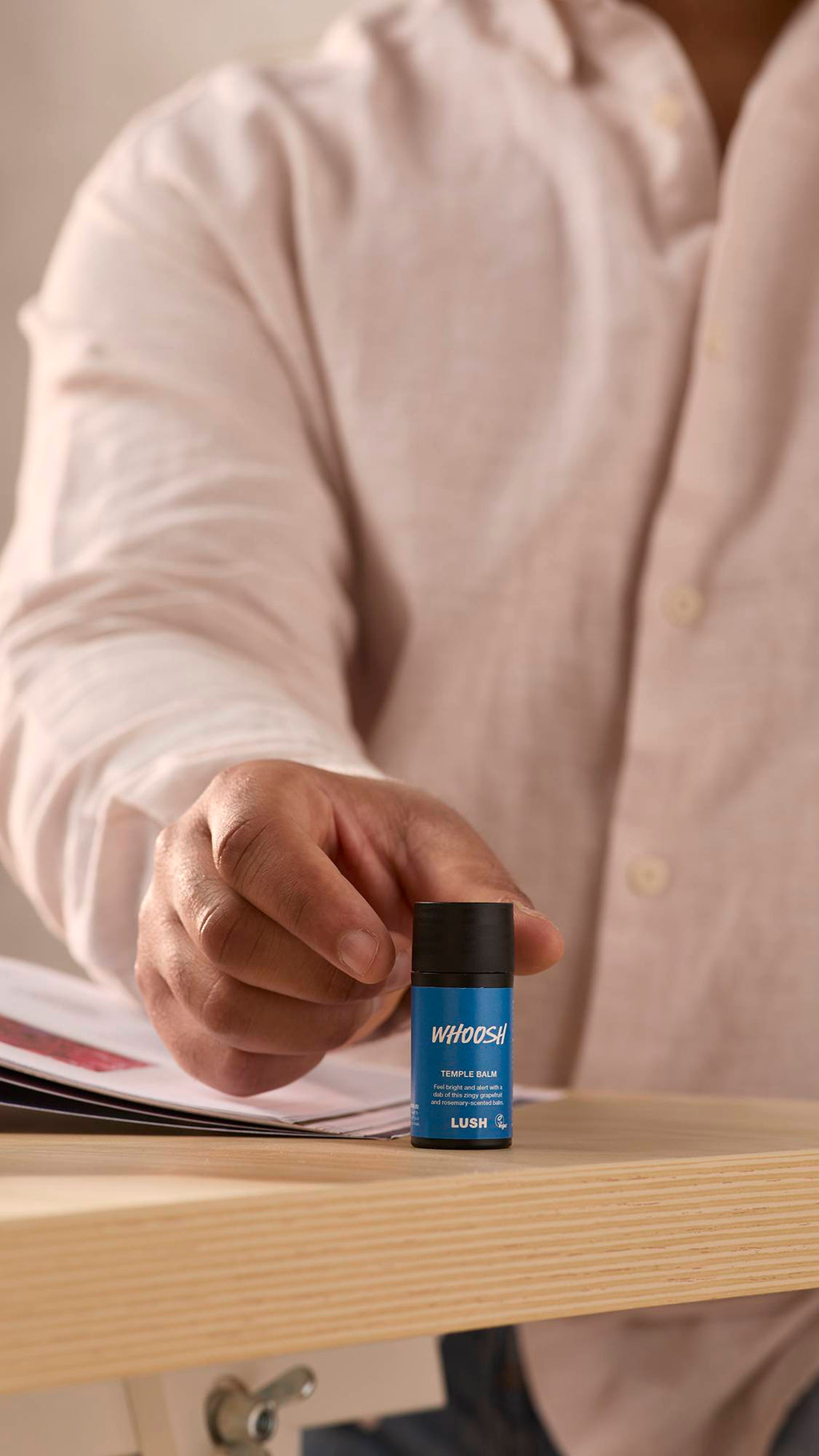 The image shows a close-up of the model sat at a desk in a shirt as they reach for the Whoosh temple balm which is sat on the desk corner facing the camera.